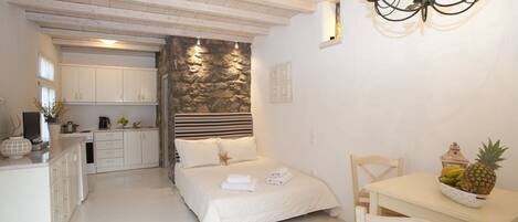 A bedroom with a bed, a bedside table with a fruit bowl, a white desk, a chair, and a kitchen area in the background.
