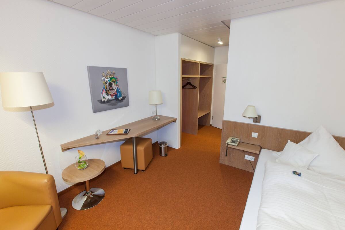 Quarto de hotel com cama, escrivaninha, cadeira, cabideiro e um quadro na parede.