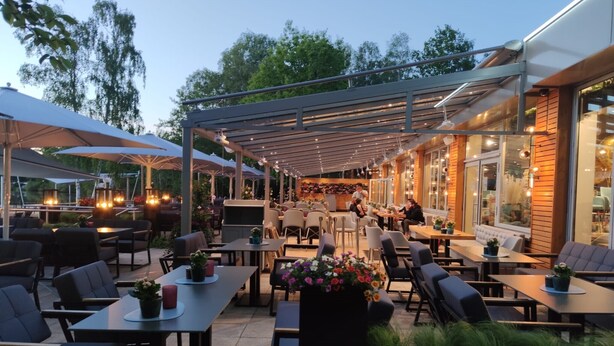 An outdoor patio area with tables and chairs, some with umbrellas. There are potted plants and a grill. - Hotel am Springhorstsee (Burgwedel)