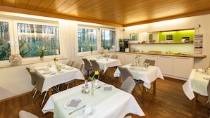 A dining area with multiple tables set for meals, large windows with a view of trees, and a kitchen area with a green-lit counter. - Hotel am Springhorstsee (Burgwedel)