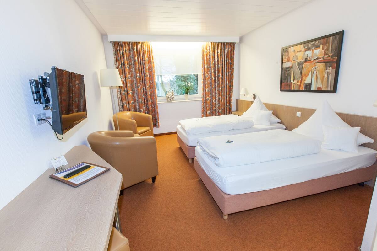 Quarto de hotel com duas camas, uma escrivaninha, uma cadeira, um quadro na parede e uma janela com cortinas.