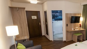 A hotel room with a wooden ceiling, a desk with a flat-screen TV, a bed, a chair, a lamp, and a door leading to a bathroom. - Hotel am Springhorstsee (Burgwedel)