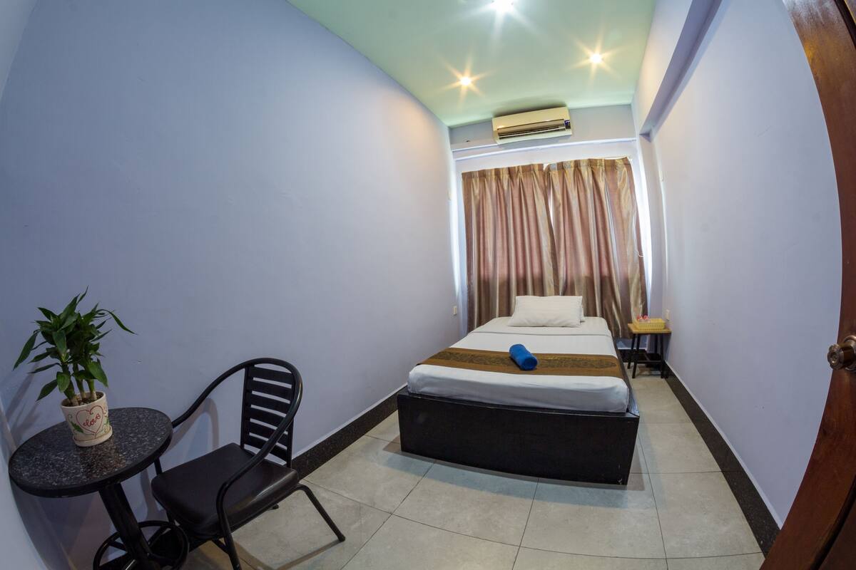 Een compacte hotelkamer met een bed, een klein tafeltje met stoel en een potplant.