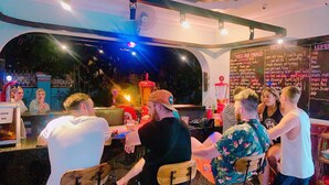 Un espace bar avec des tables et des chaises, un tableau noir où sont écrites des phrases, et un grand ventilateur.