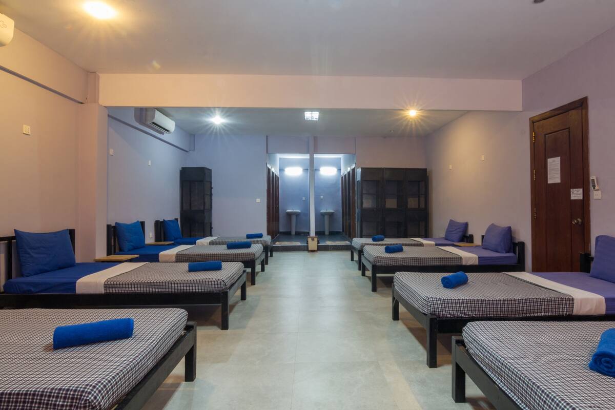 Een hotelkamer met vier enkele bedden, elk met een blauw kussen en een geruit sprei. Er is een spiegel aan de muur, een houten deur en een klein tafeltje in de hoek.