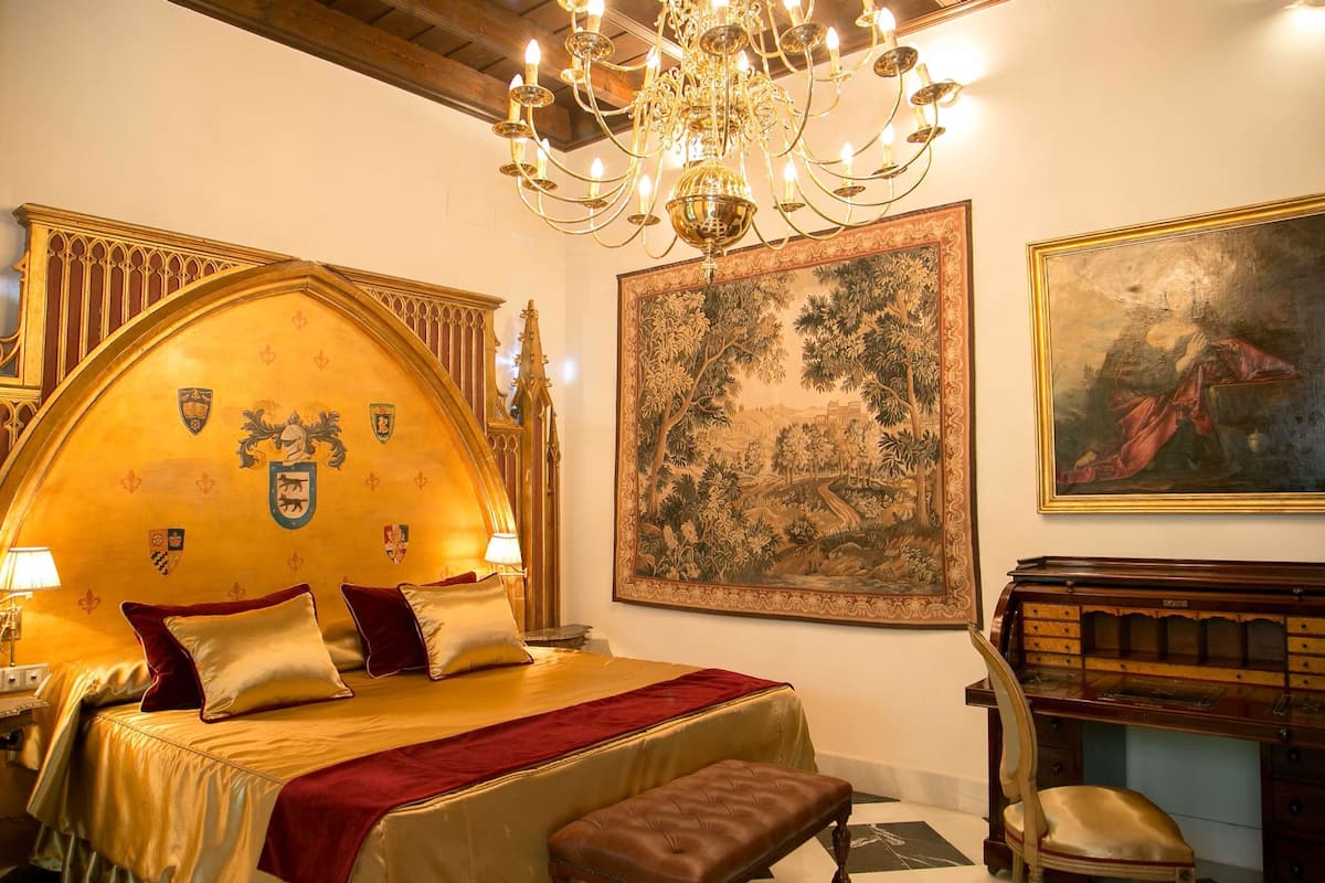 A luxurious bedroom with a large bed, a chandelier, a large tapestry, and a piano.