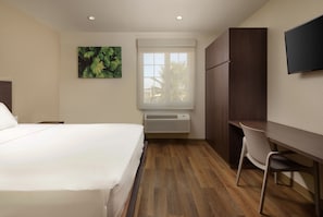 A hotel room with a bed, a desk, a chair, a window with blinds, a wall-mounted TV, and a painting.