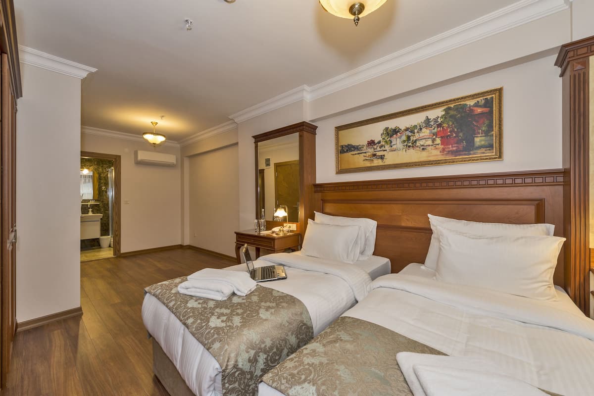 A hotel room with two beds, a desk with a laptop, a painting on the wall, and a bathroom visible through an open door.