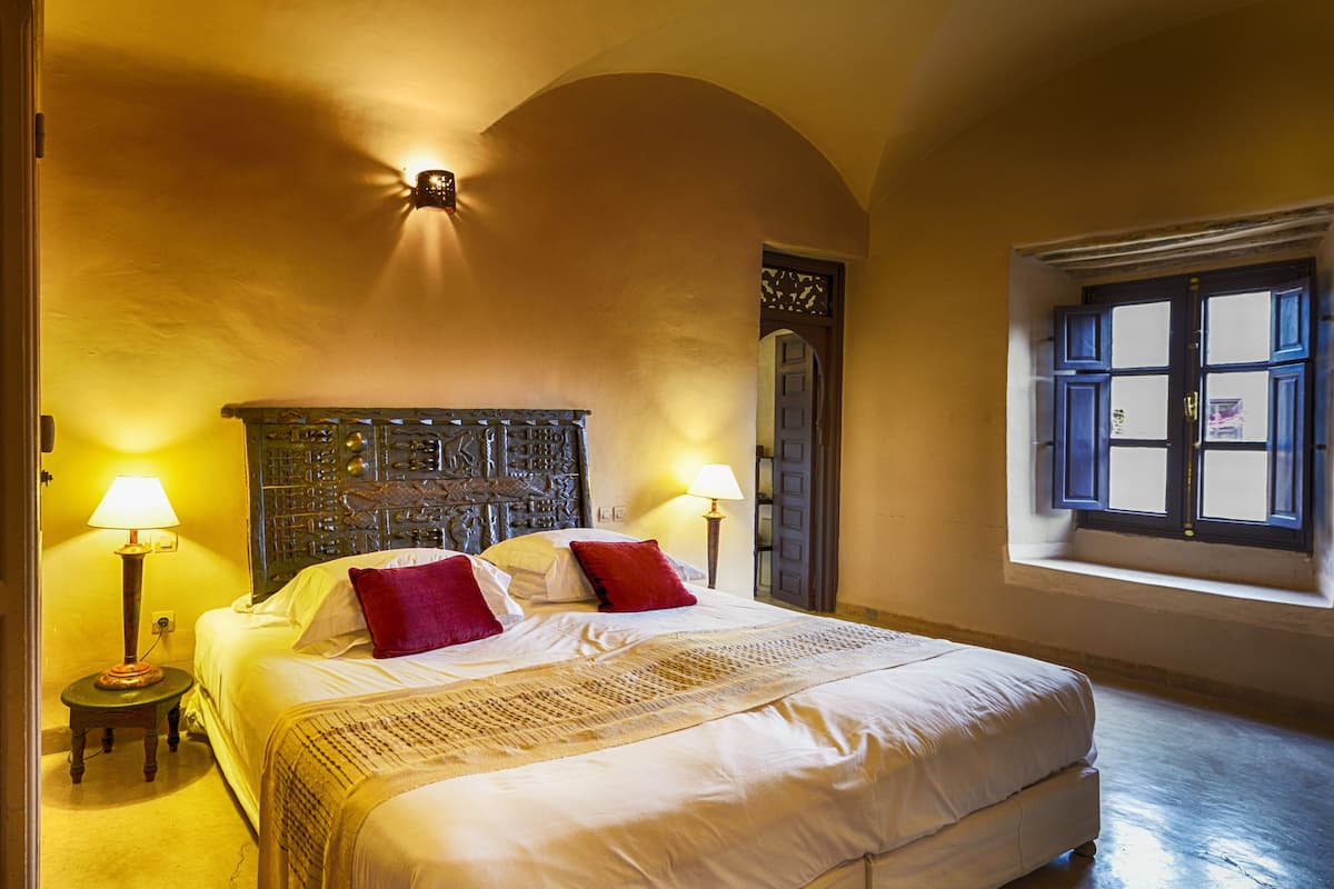 A bedroom with a large bed, two bedside lamps, a decorative headboard, and a window with a view.