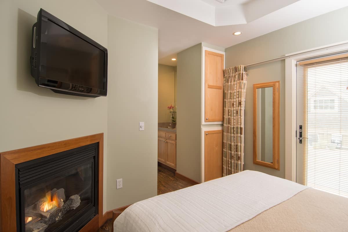 A bedroom with a fireplace, a television mounted on the wall, a bed, and a view of the kitchen and bathroom through an open door.