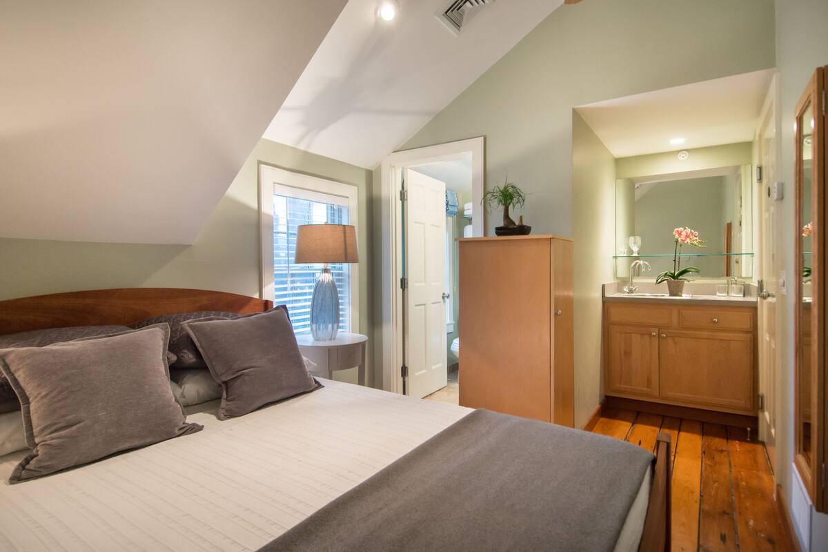 A bedroom with a bed, pillows, a bedside table, a lamp, a wooden wardrobe, and a bathroom visible through an open door.