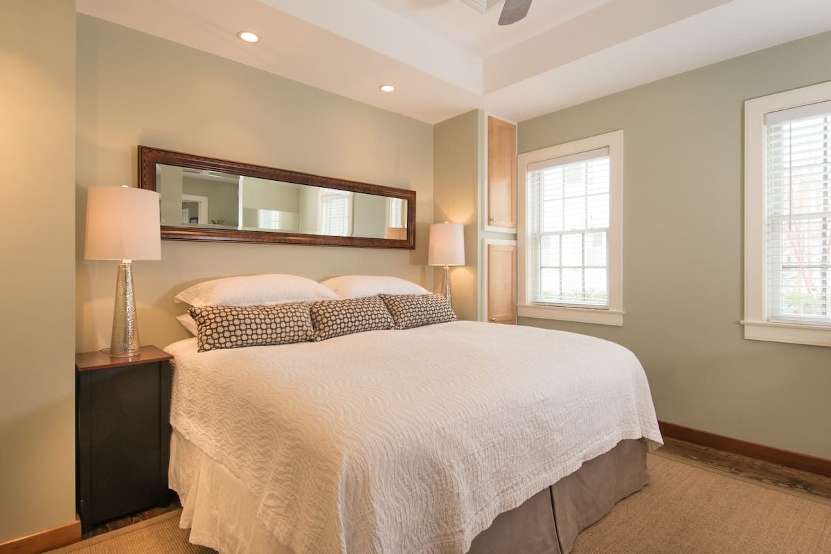 A neatly arranged bedroom with a large bed, two bedside tables with lamps, a mirror above the bed, and a window with blinds.