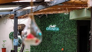 An outdoor seating area with tables and chairs, a green wall with a 'Cold Drinks' sign, and hanging lights.