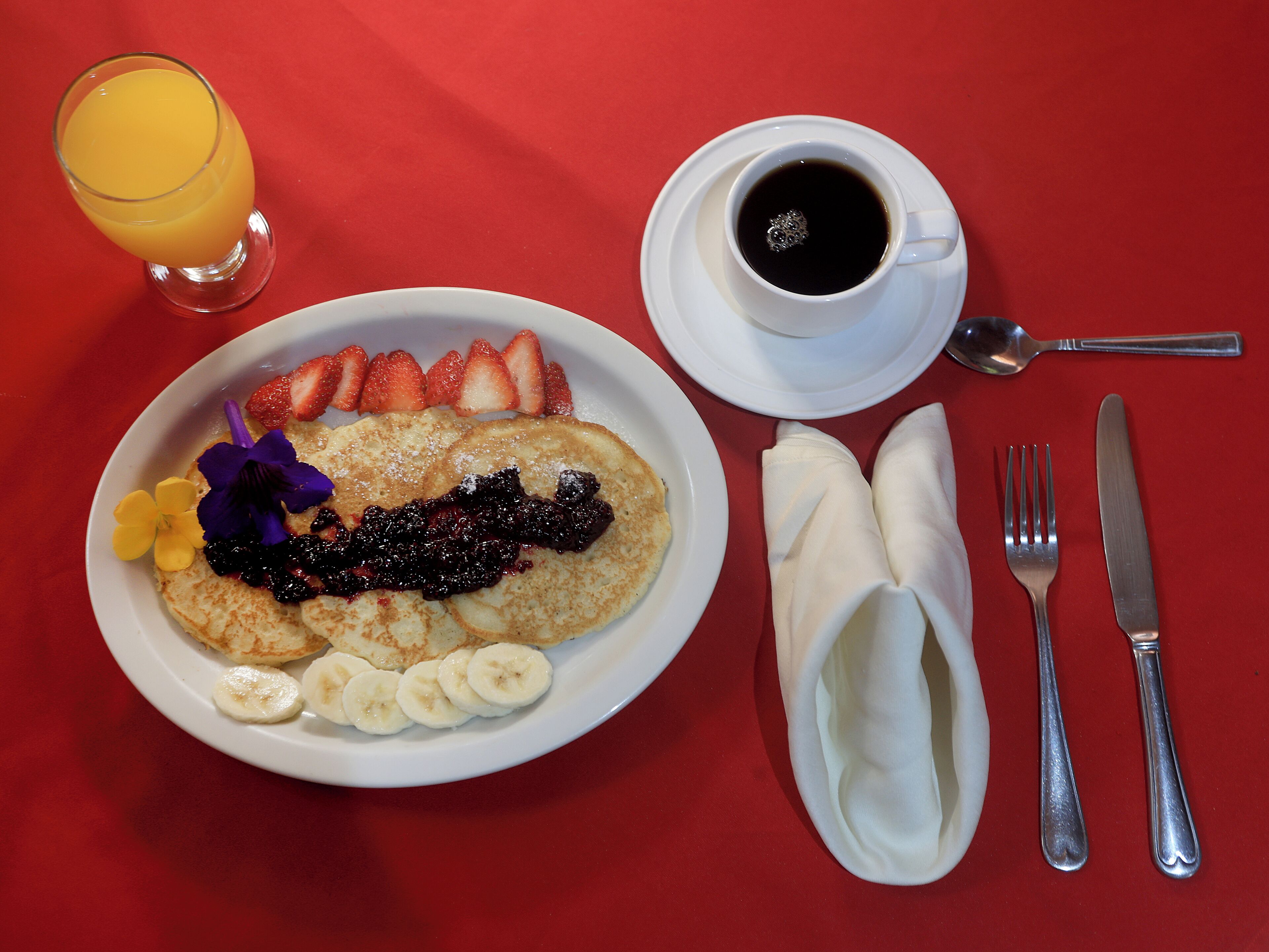 A breakfast plate with pancakes, strawberries, bananas, and a cup of coffee on a red table.