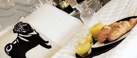 A neatly made bed with a pillow featuring a black and white dog silhouette, a tray with croissants and orange juice, and a bedside table with a bottle and a glass.