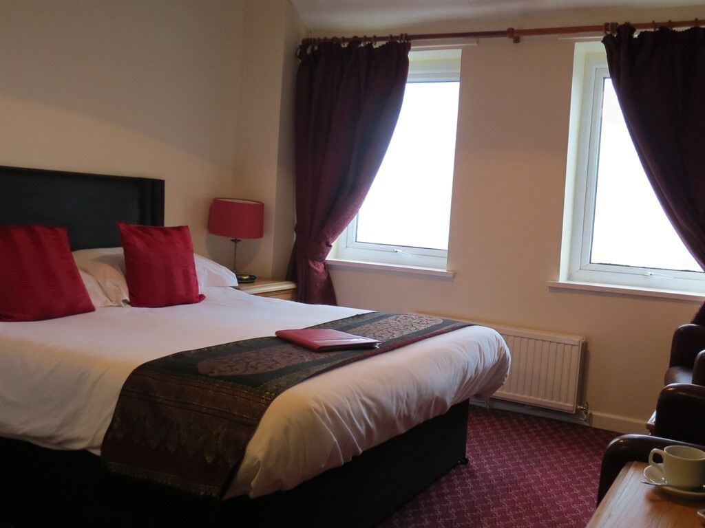 A hotel room with a large bed, two windows with curtains, a bedside table with a lamp, and a coffee table.