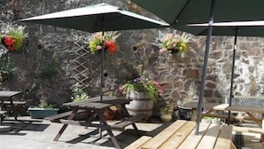 An outdoor patio with wooden tables and benches, stone walls, and hanging flower baskets. - The Wyche Inn (Malvern)