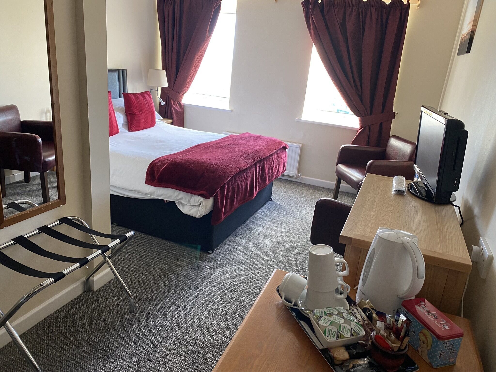 A hotel room with a bed, a desk with a computer, a chair, a bedside table with a cup and a tray of breakfast items, and a window with curtains.