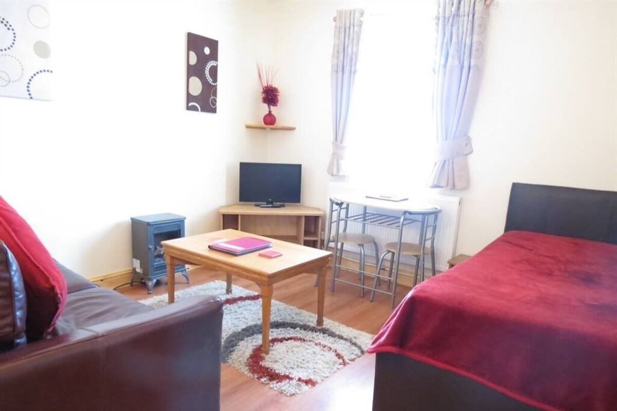 A small, well-lit room with a sofa, a bed, a desk with a computer, and a small table.