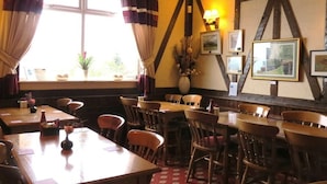 A restaurant with wooden tables and chairs, a large window with curtains, and wall-mounted picture frames. - The Wyche Inn (Malvern)