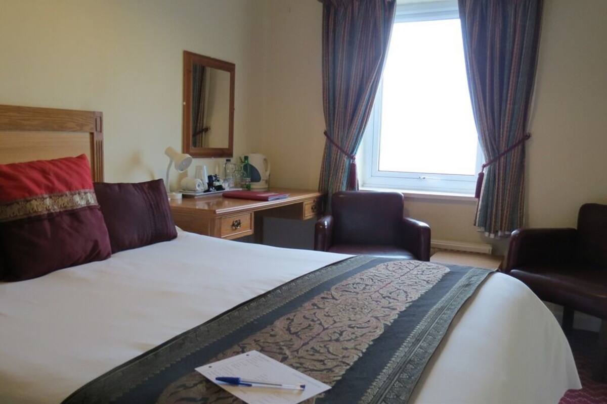 A hotel room with a large bed, a desk, two chairs, a window with curtains, and a mirror on the wall.