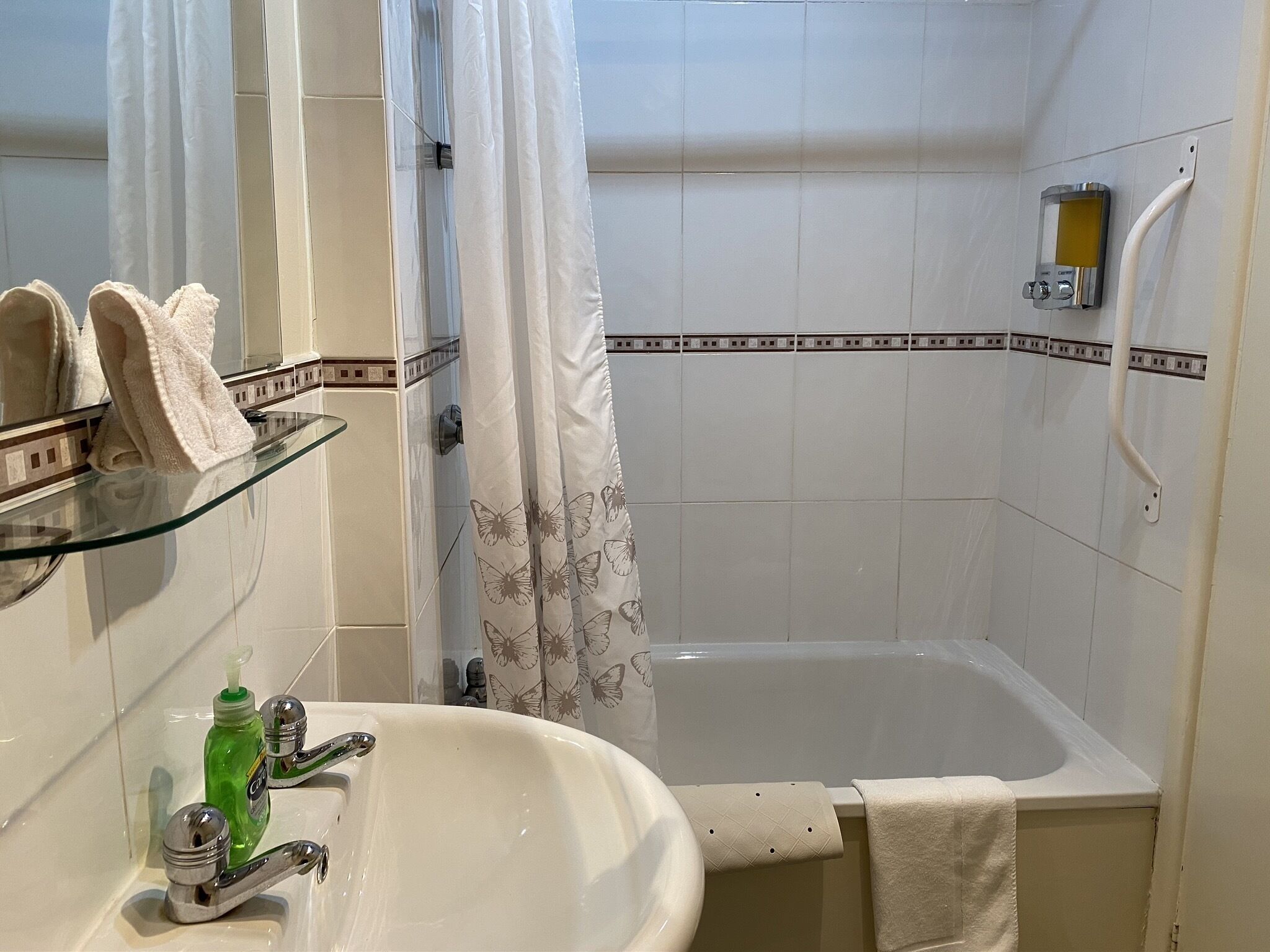 Un baño con repisas de vidrio que sostienen toallas, un lavamanos con una botella verde y una zona de ducha con cortina.