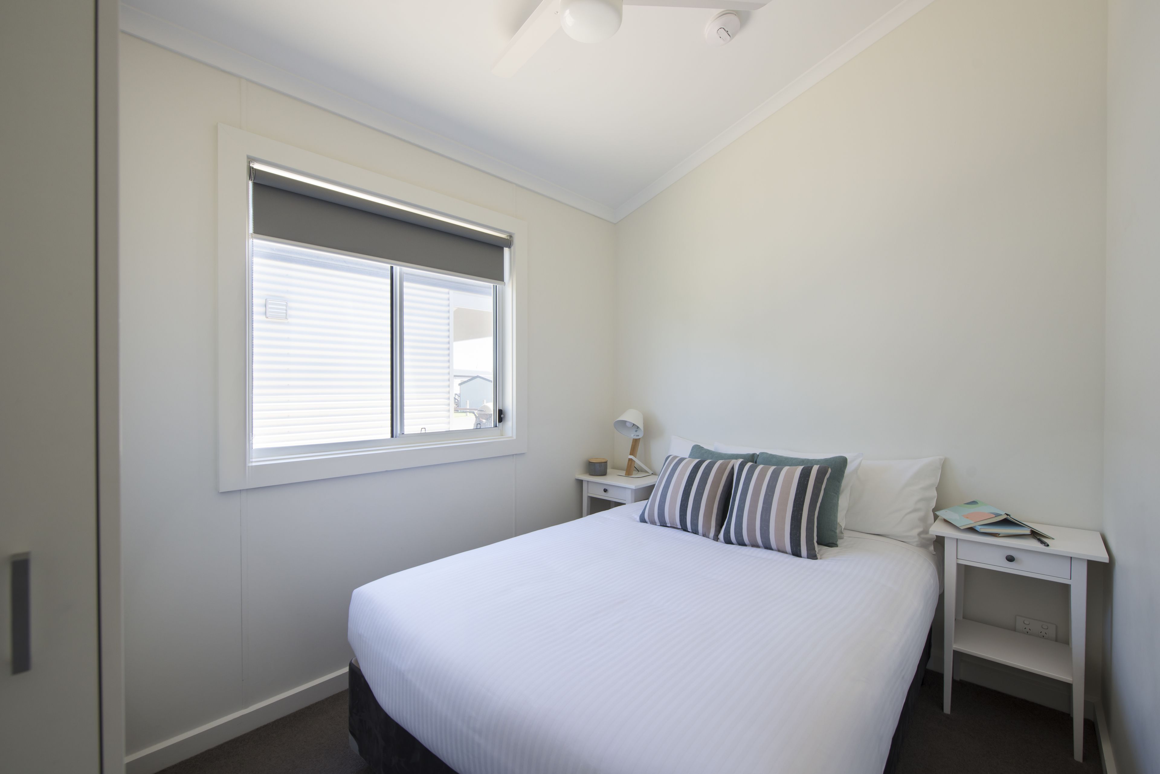 A bedroom with a large bed, a window with blinds, a bedside table with a lamp, and a nightstand.
