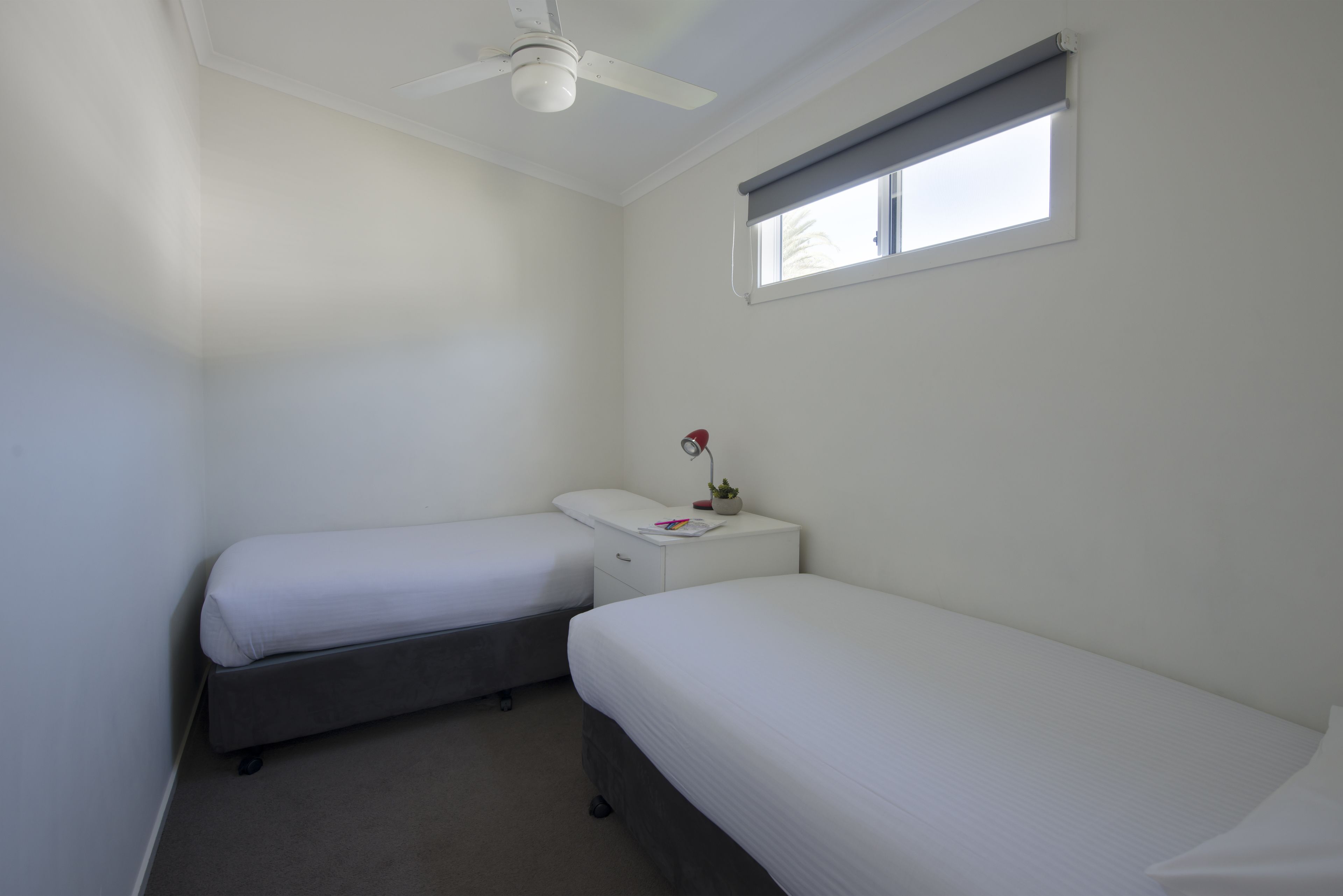 A small, clean room with two single beds, a ceiling fan, a window with a blind, and a bedside table with a lamp and a small plant.