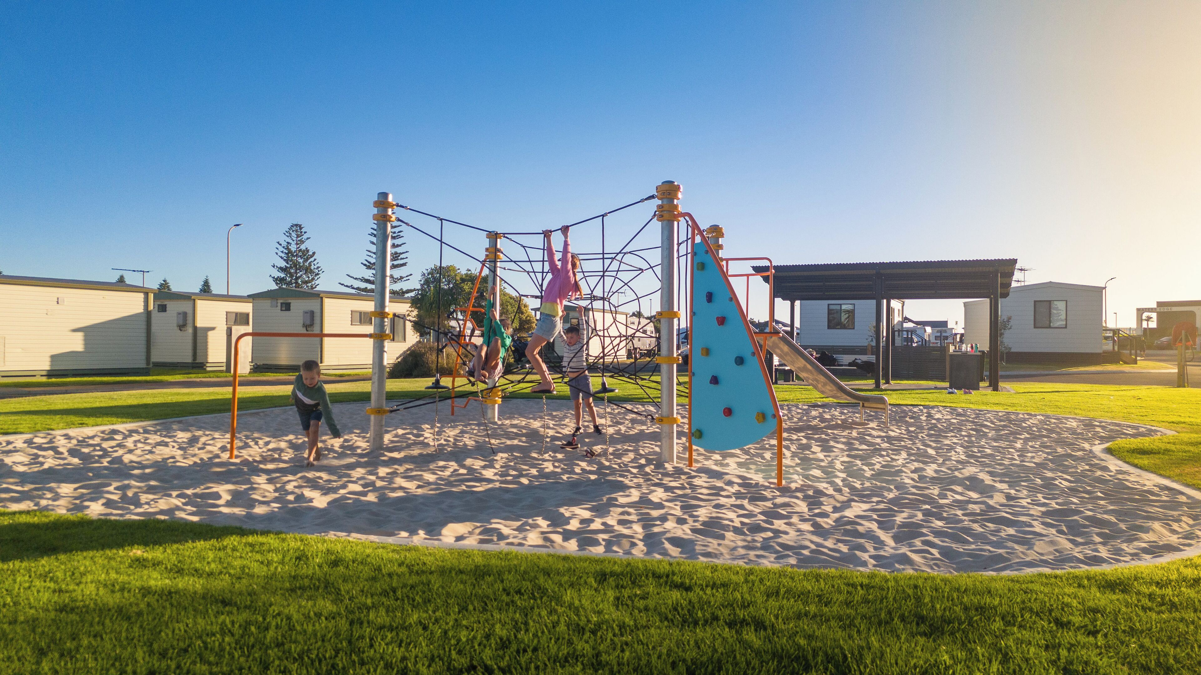 A playground with climbing structures and a slide, surrounded by a sandy area and grass.