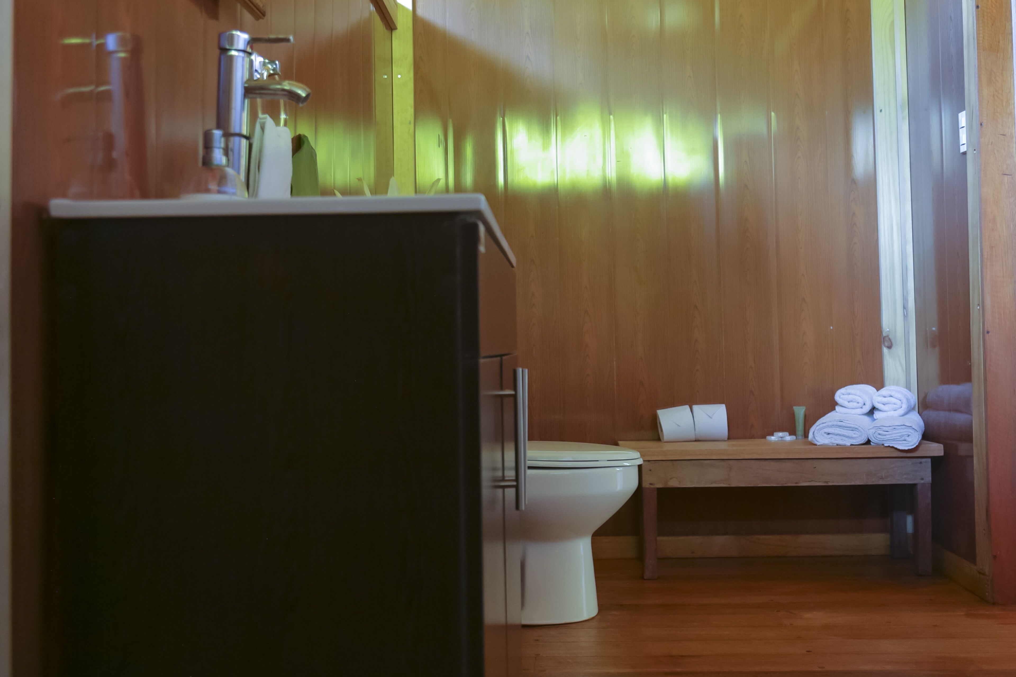 A wooden-paneled bathroom with a toilet, a sink, and a small table with towels.