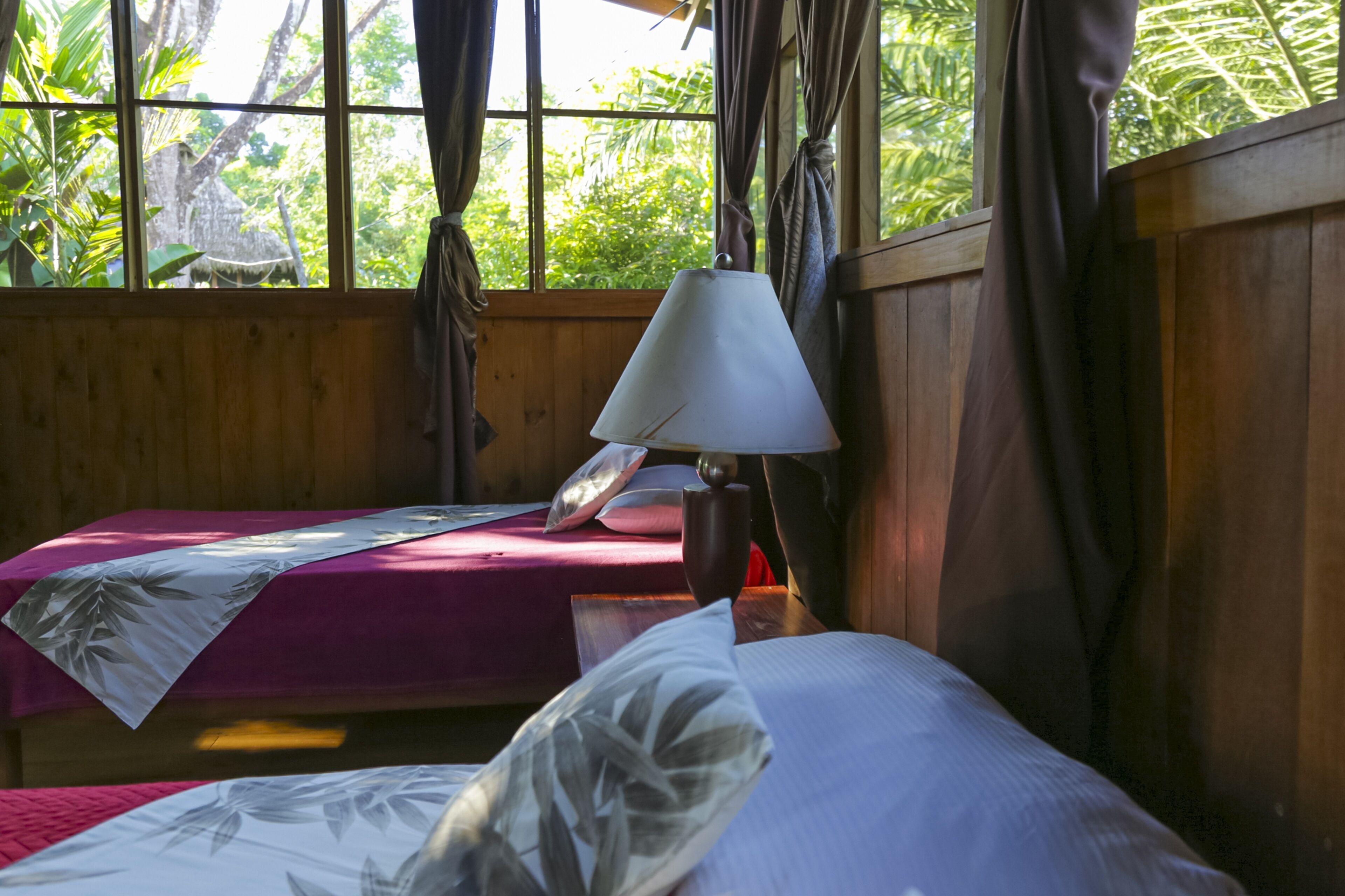 Una habitación con dos camas, un muro de madera, una ventana con cortinas y una lámpara de mesa.