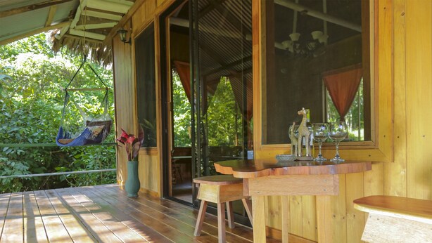 Une terrasse en bois avec un hamac suspendu, une table en bois avec des chaises et une fenêtre avec des rideaux.
