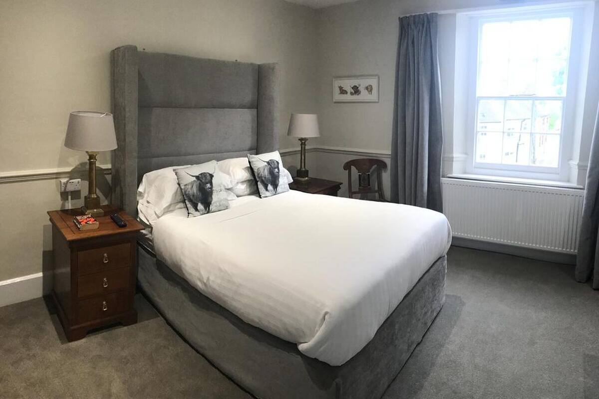 Um quarto bem arrumado com uma cama grande, duas mesas de cabeceira com luminárias, uma cadeira, uma janela com cortinas e um quadro na parede.