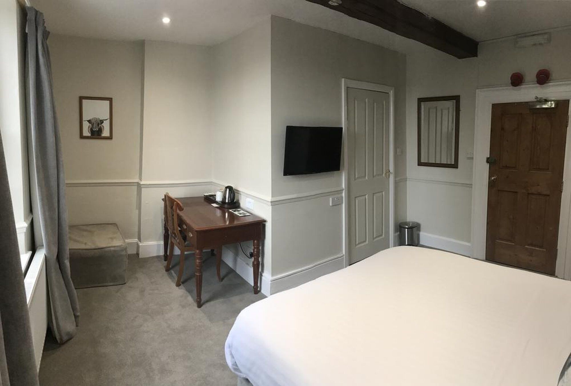A hotel room with a bed, a desk with a chair, a television mounted on the wall, a door, and a small trash can.