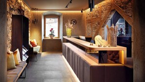 A modern reception area with a stone wall, a wooden reception desk, a bench with cushions, and a window with a view of the city. - Gasthof Hotel Post (Strass im Zillertal)