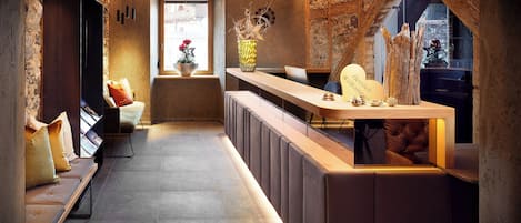 Un área de recepción moderna con pared de piedra, mostrador de recepción de madera, un banco con cojines y una ventana con vista a la ciudad.