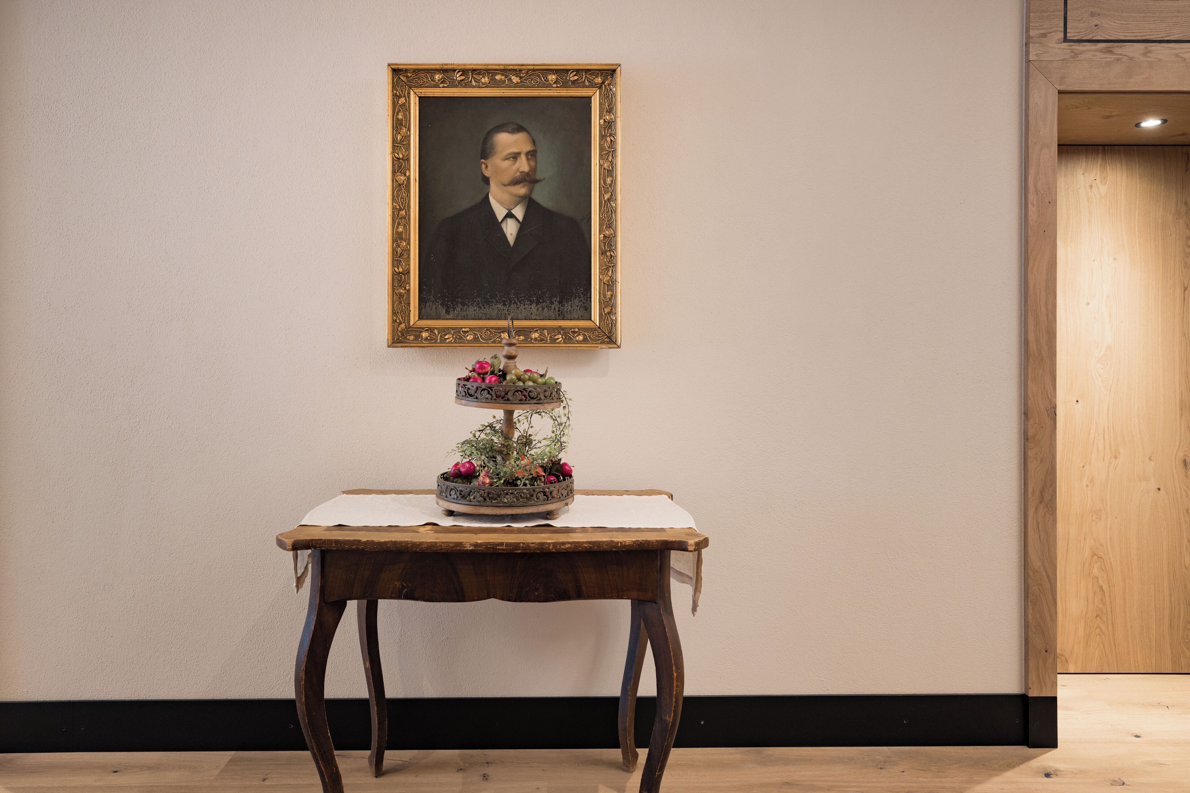 Un retrato enmarco en una pared, una pequeña mesa con un arreglo floral y suelo de madera.