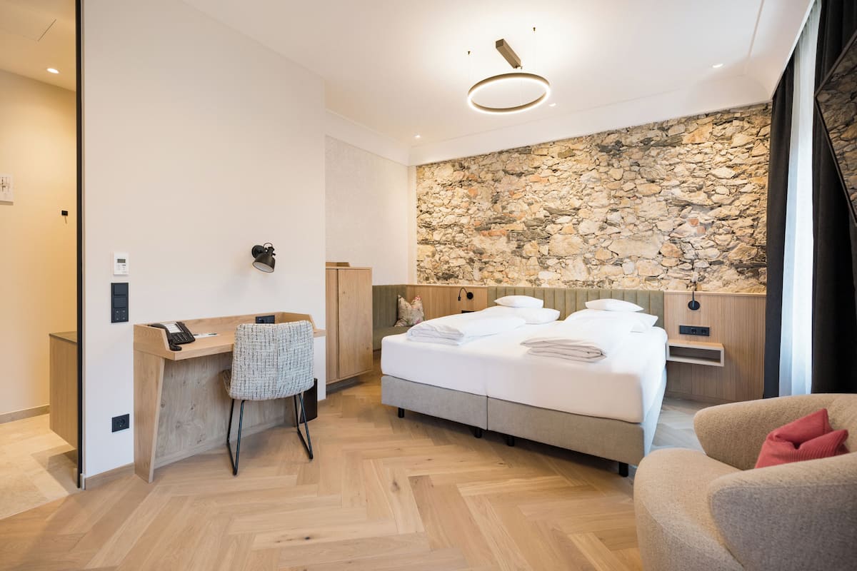 Ein modernes Hotelzimmer mit einem Holztisch, einem Bett mit weißer Bettwäsche, einer Steinwand und einem großen Fenster.