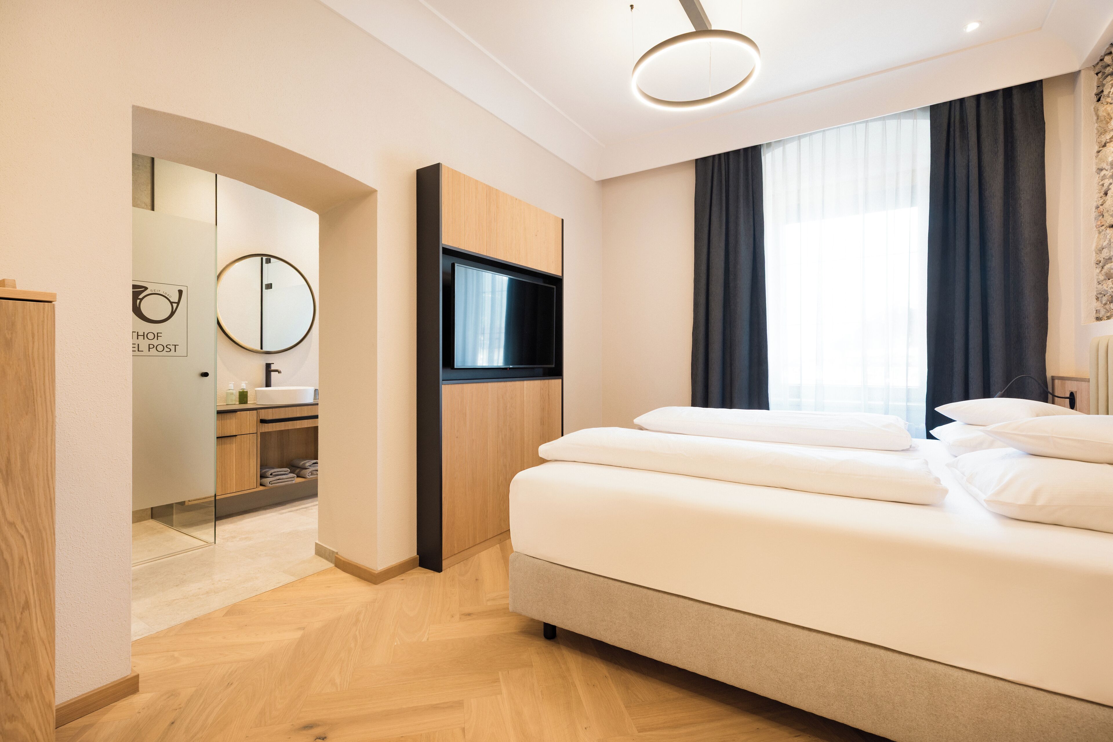 A modern hotel room with a large bed, a flat-screen TV, a mirror, and a bathroom visible through an archway.
