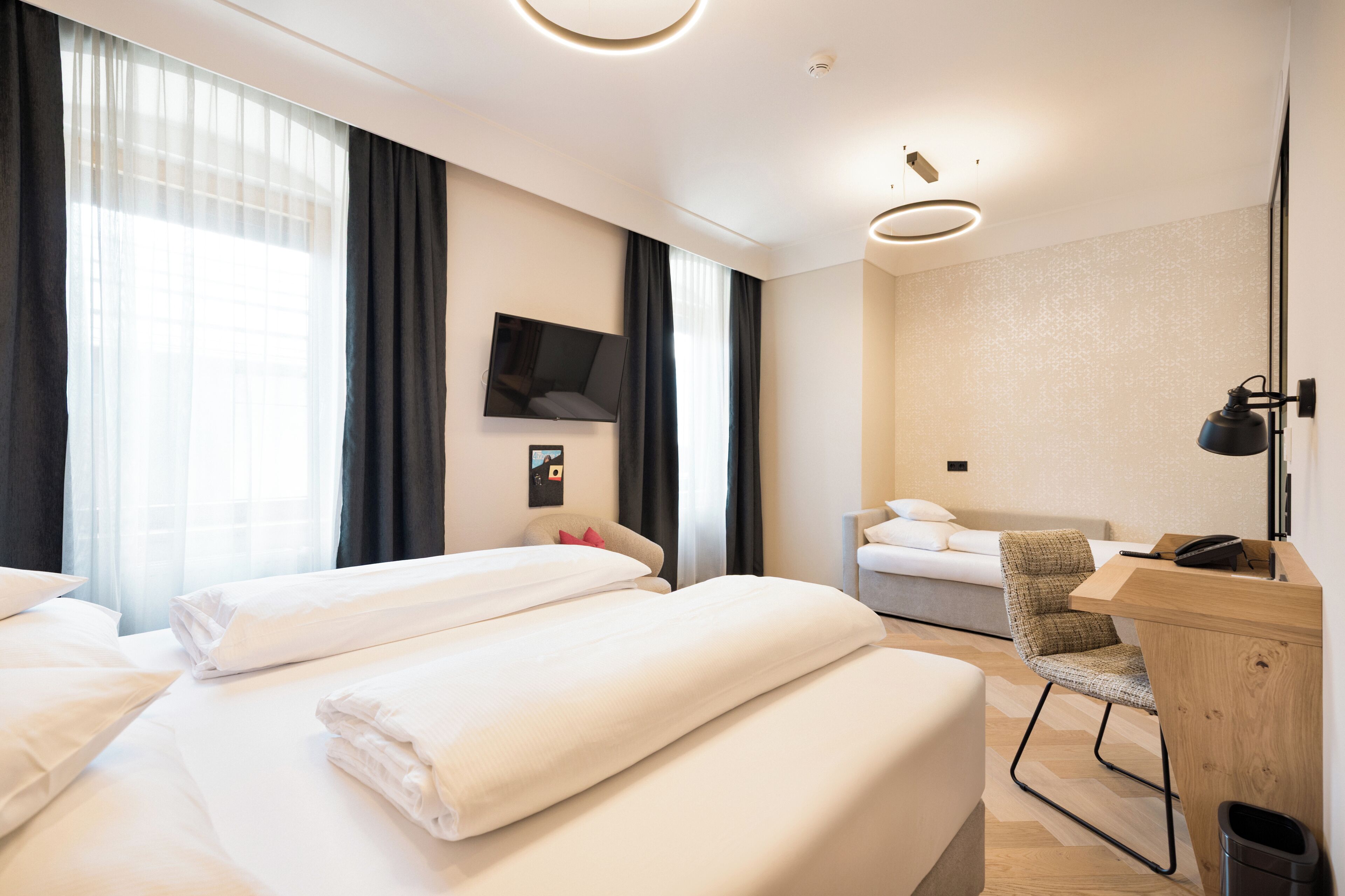 A hotel room with two beds, a desk, a chair, a TV, and a large window with curtains.