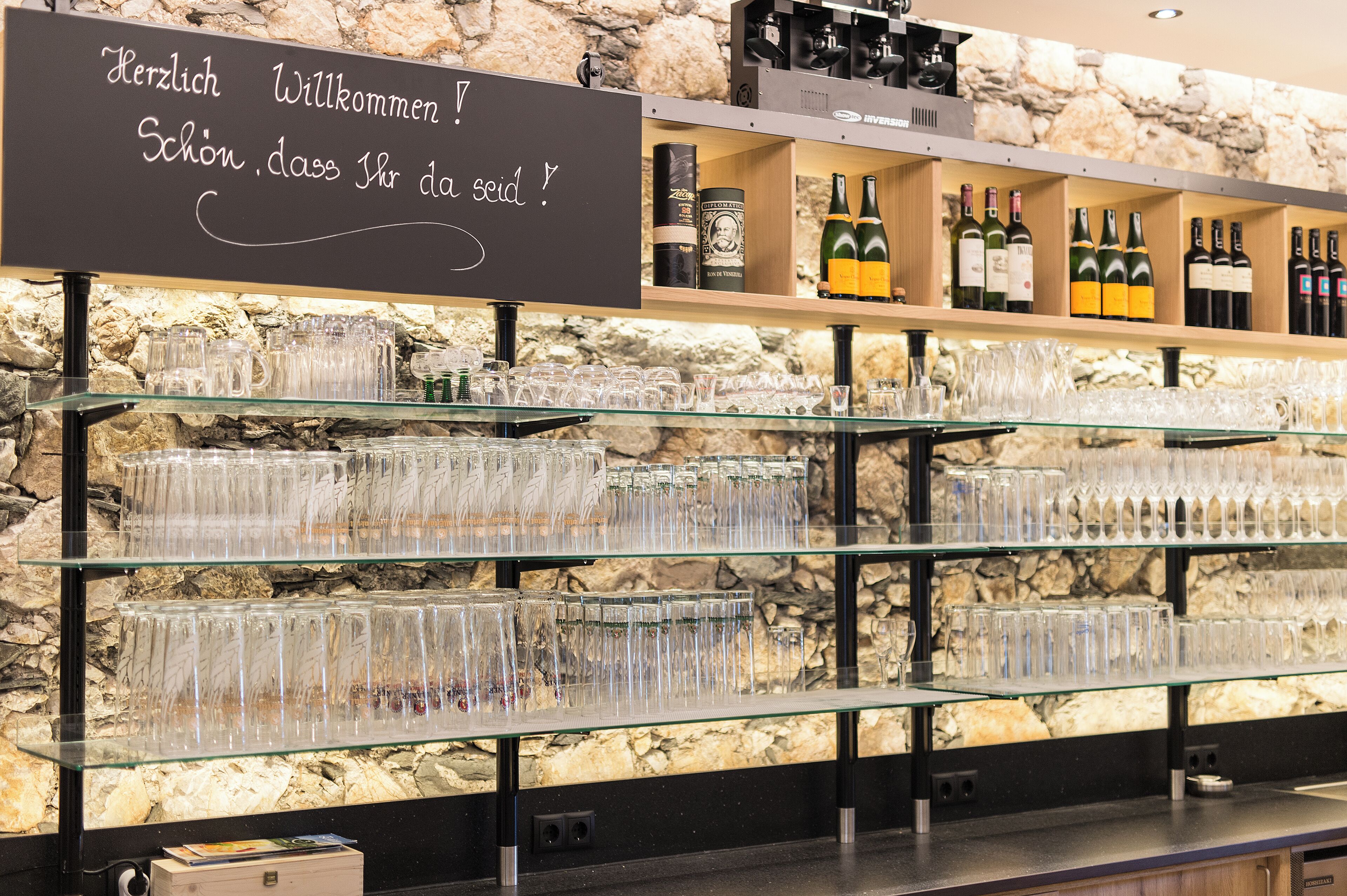 A bar area with a stone wall background, shelves with wine bottles, and a sign with a greeting in German.