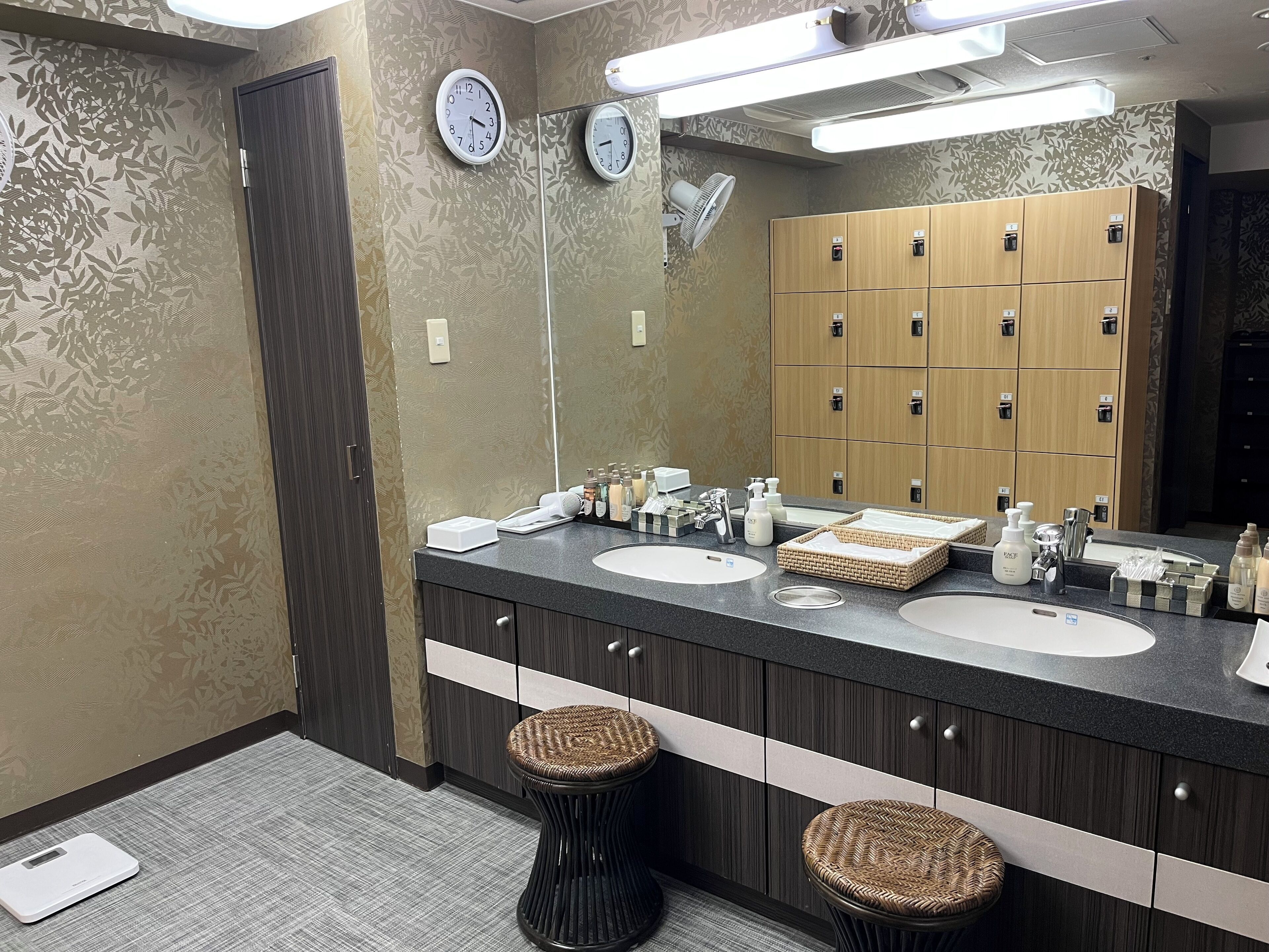 Un baño moderno con dos lavamanos, un espejo grande, relojes de pared y una fila de taquillas de madera.