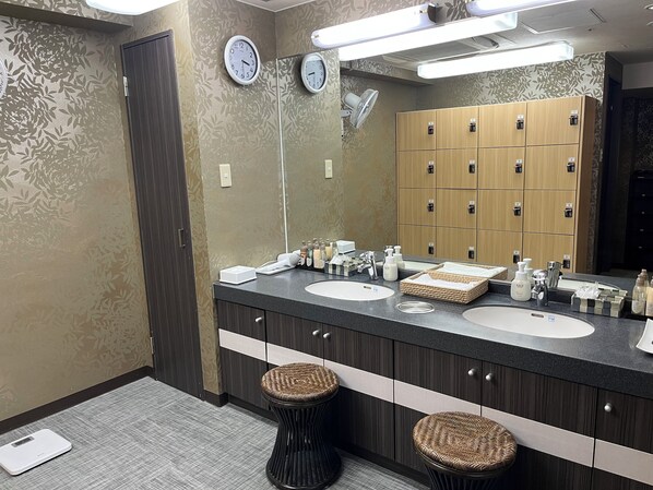 Un baño moderno con dos lavamanos, un espejo grande, relojes de pared y una fila de taquillas de madera.