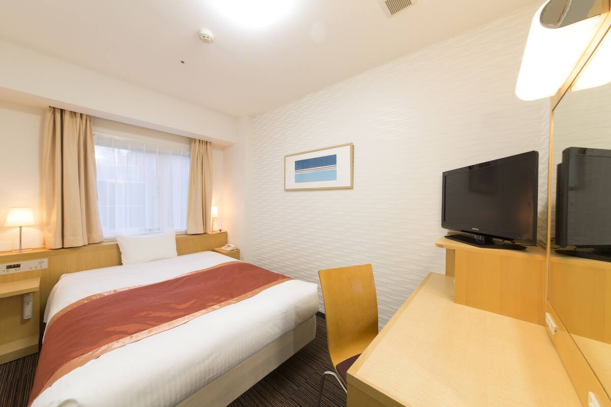 A hotel room with a bed, a desk, a chair, a television, a nightstand, a lamp, and a window with curtains.