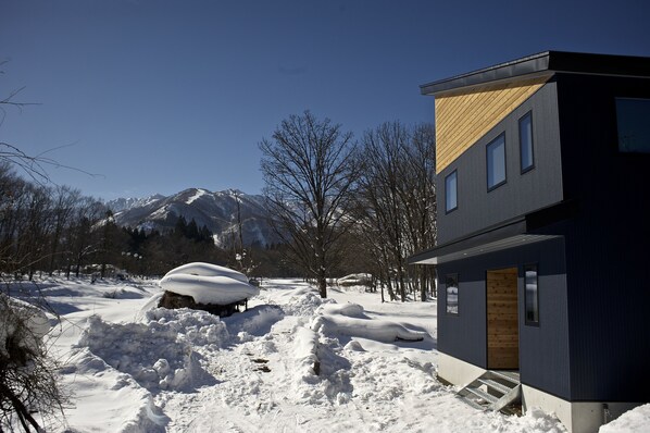 Une maison moderne à deux étages, dotée d’une partie du toit en bois, entourée d’un paysage enneigé et de montagnes.