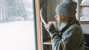 A person in a knit hat and sweater sitting by a window with a view of a snowy landscape. - Wagaya (Hakuba)
