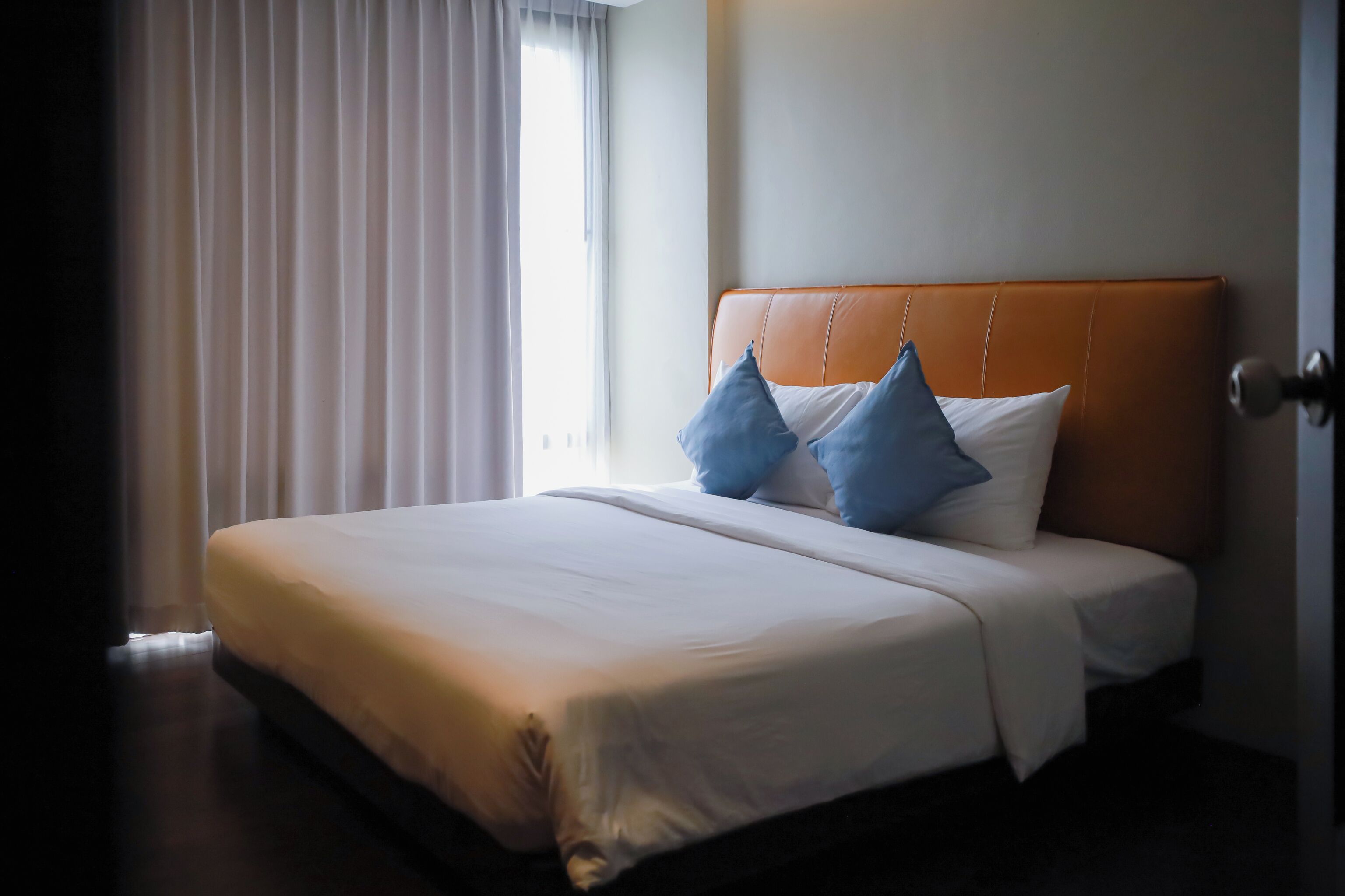 A neatly made bed with white linens and two pillows, a brown headboard, and a window with white curtains.