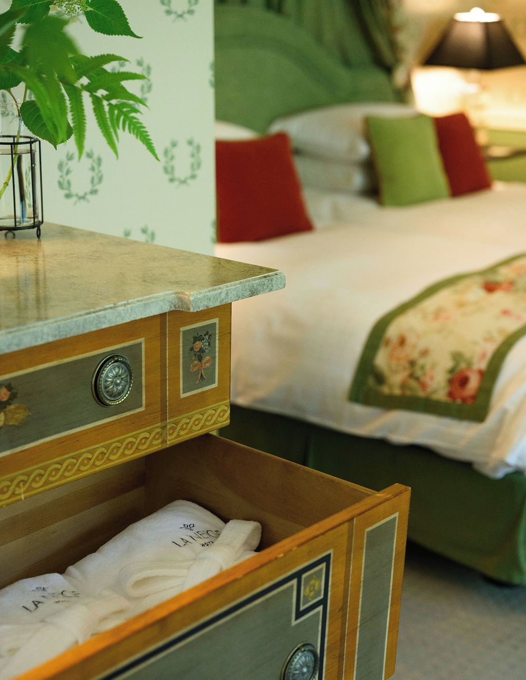 A wooden dresser with drawers, a bed with a floral bedspread, and a green wall with a plant.