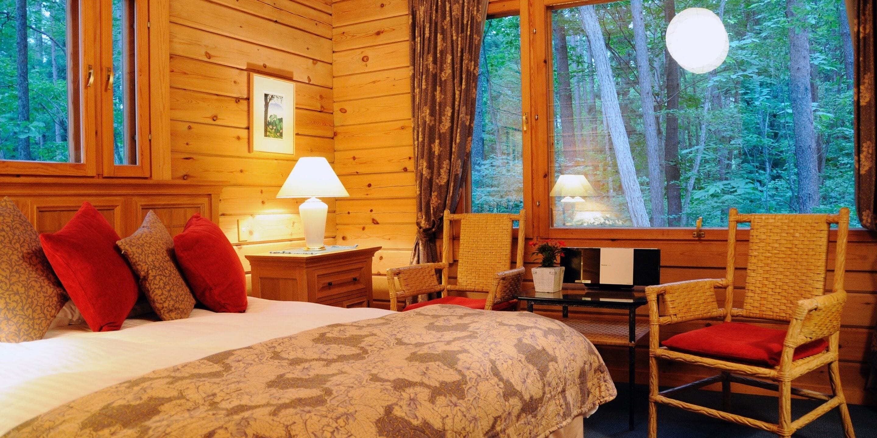 Acogedora habitación tipo cabaña de madera con cama, dos sillones, una mesa y ventana con vista al bosque.
