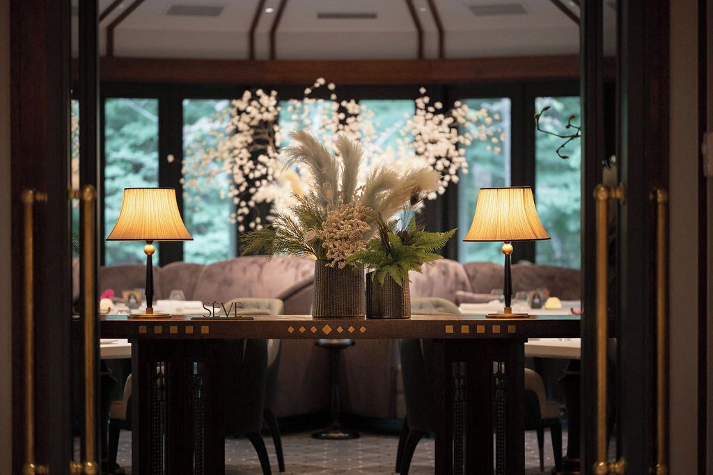 A dining area with a table set for two, flanked by two lamps and a vase with dried flowers.